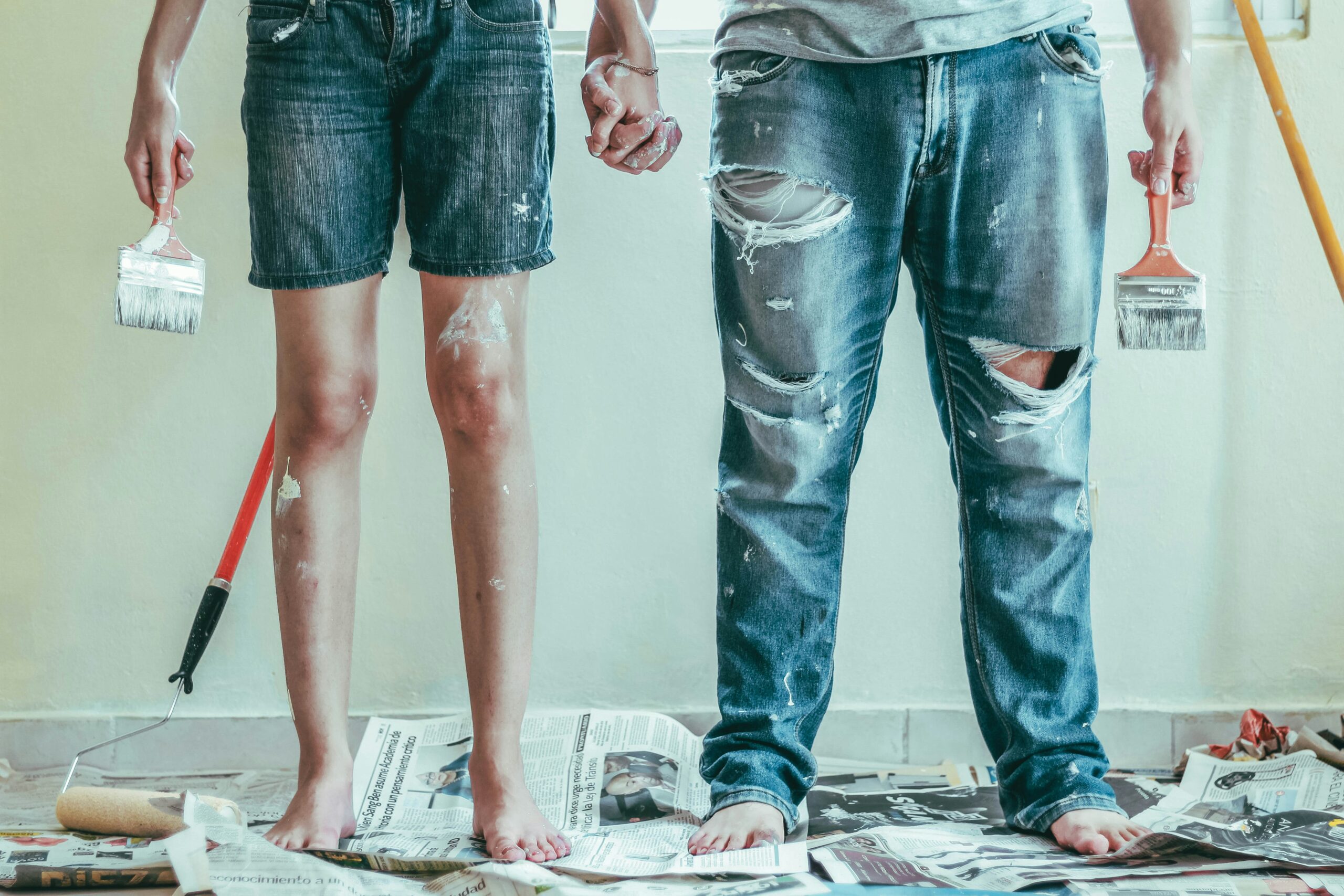 Shot of the legs of a couple holding paintbrushes with paint-stained clothes